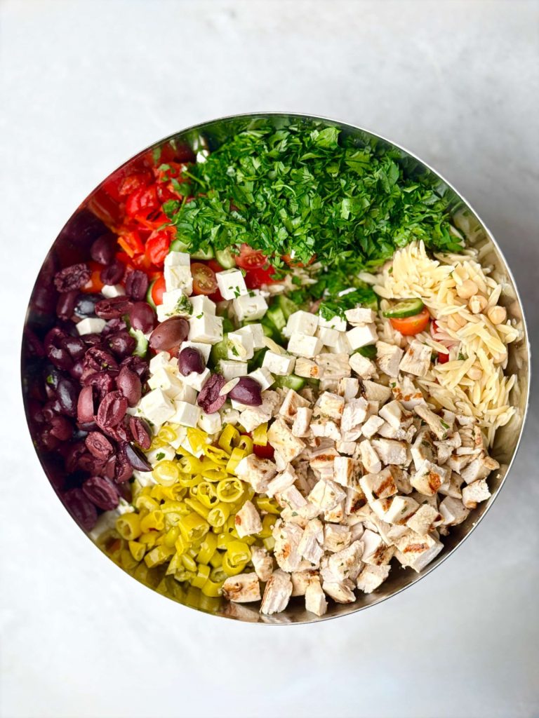 Large mixing bowl filled with the colorful ingredients for Greek chicken and orzo pasta salad, including chopped chicken, briny olives, cherry tomatoes, cucumbers, parsley, feta, and orzo.