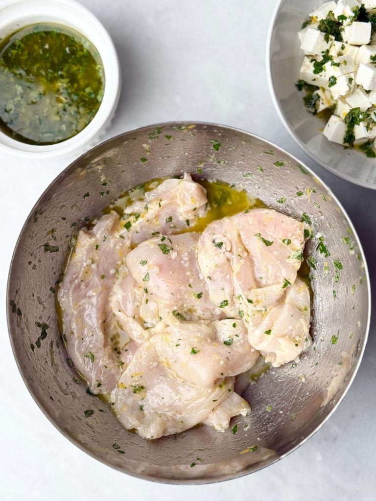 Raw skinless chicken breasts marinating in a lemon herb dressing beside a small bowl of feta cheese cubes in Greek dressing, prep stage for Greek chicken and orzo pasta salad.