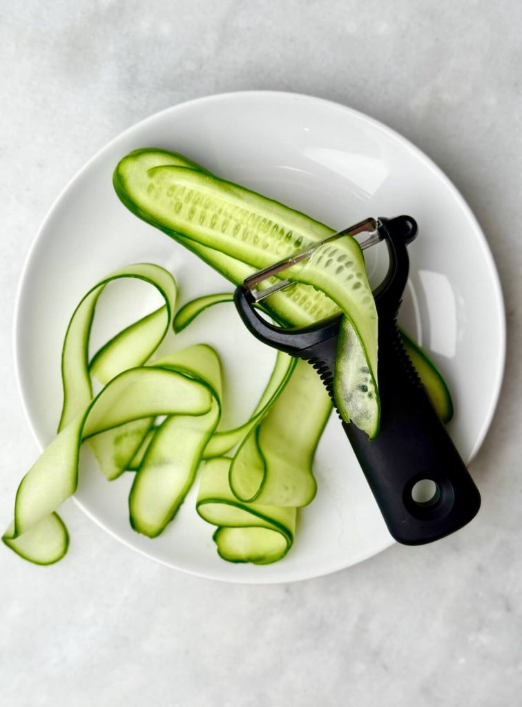 Thin cucumber ribbons on a white plate with a vegetable peeler, ready for a fresh summer watermelon salad.