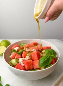 Chili-lime dressing being poured over a colorful Watermelon Feta Salad with Cucumber, basil, and creamy feta in a wide white bowl.
