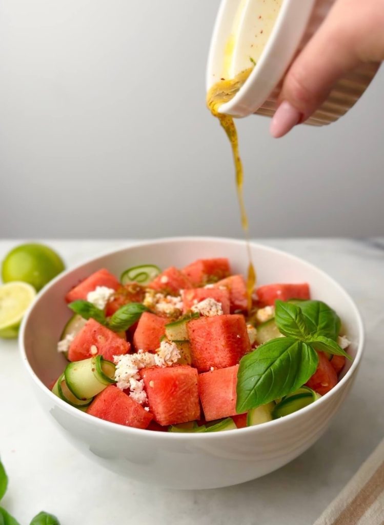 Chili-lime dressing being poured over a colorful Watermelon Feta Salad with Cucumber, basil, and creamy feta in a wide white bowl.