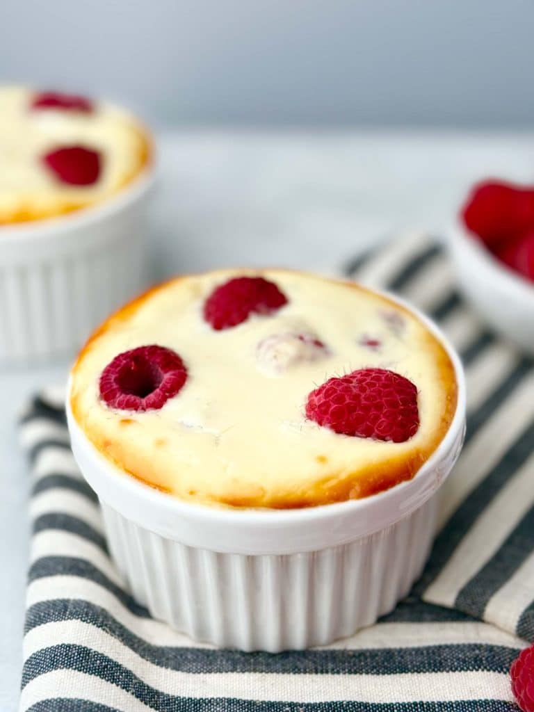 Side view of raspberry yogurt bake in white ramekin, topped with baked-in raspberries on striped cloth background.