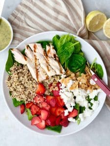 Large white bowl of spinach strawberry salad with grilled chicken, quinoa, goat cheese, sliced almonds, and a cup of poppyseed dressing on the side.