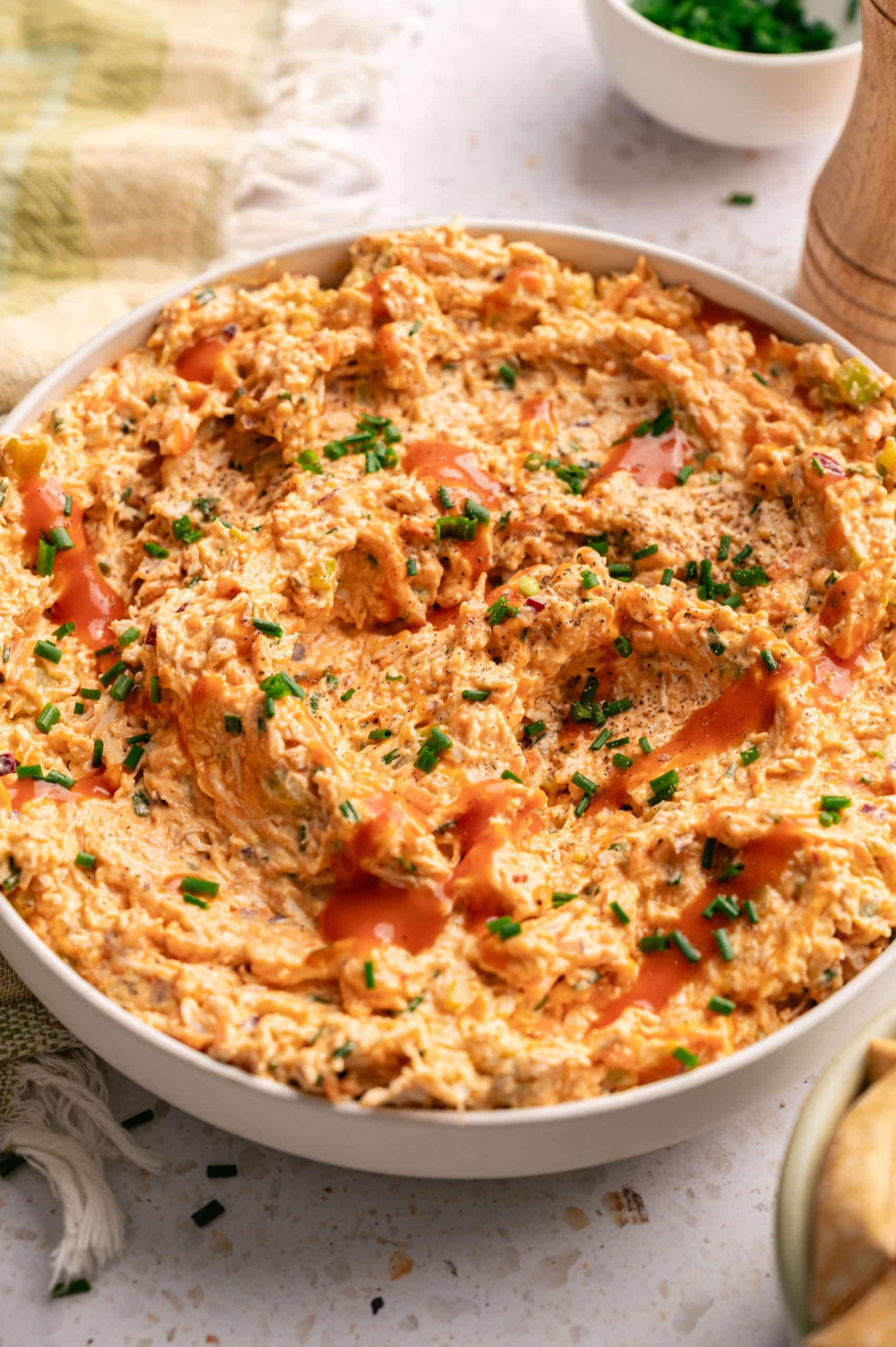 Buffalo chicken salad in a white serving bowl drizzled with hot sauce and topped with fresh chives.