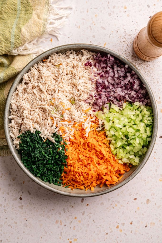 Shredded chicken, diced red onion, celery, carrots, and chives arranged in a bowl before mixing.