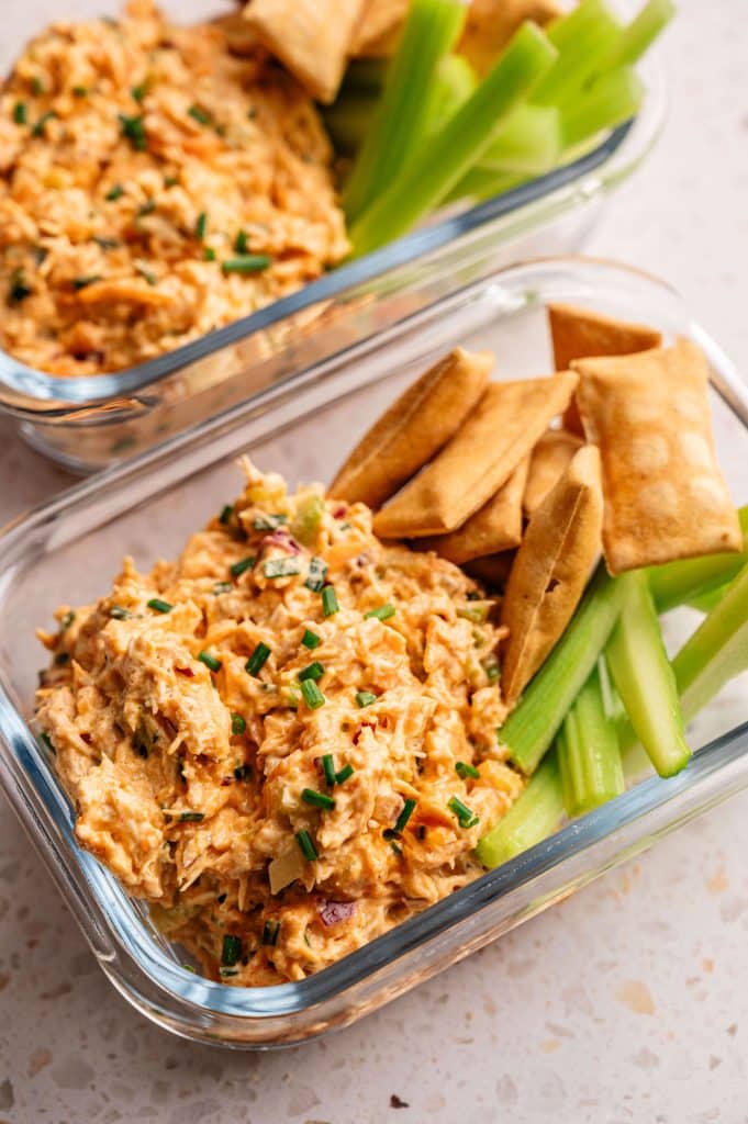 Two glass meal prep containers filled with buffalo chicken salad, pita chips, and celery sticks.