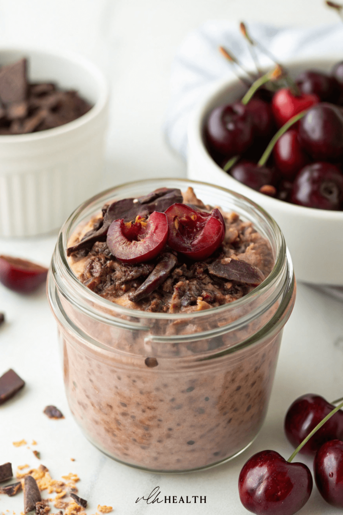 Glass jar filled with chocolate overnight oats topped with halved fresh cherries and dark chocolate chunks, surrounded by whole cherries.