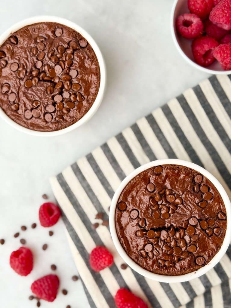 Overhead view of two white ramekins filled with chocolate protein baked oats with Greek yogurt, surrounded by fresh raspberries and mini chocolate chips.