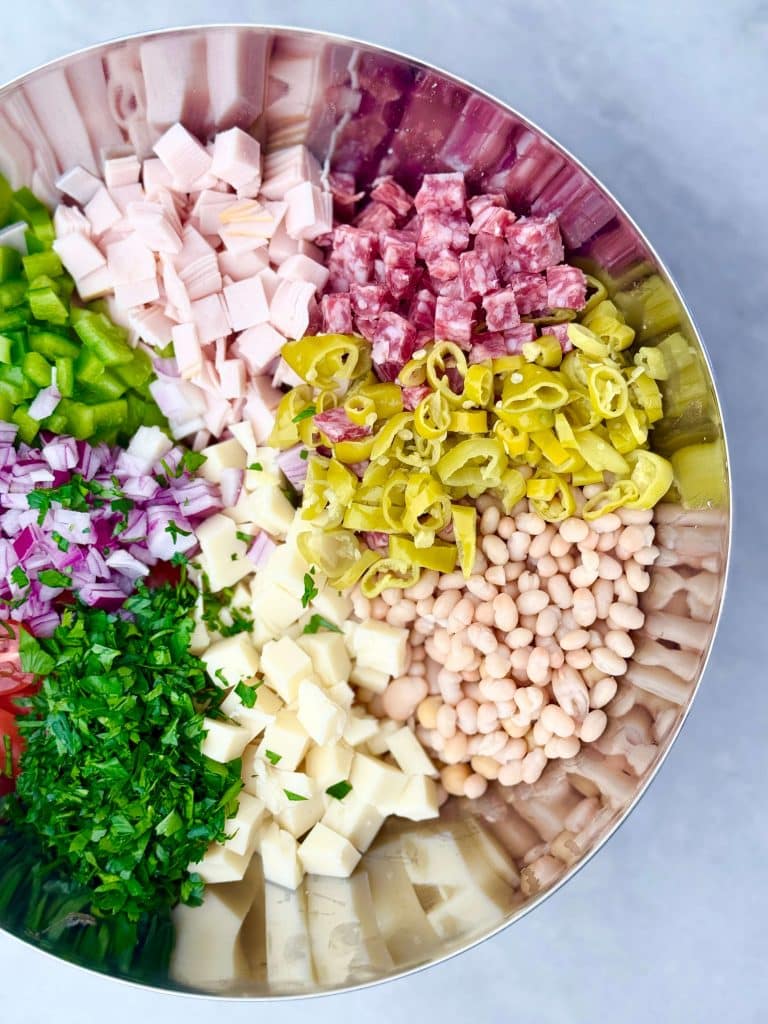 Unmixed ingredients for Italian grinder bean salad arranged in sections in a stainless steel bowl, including chopped turkey, salami, peppers, onion, parsley, cheese, and beans.