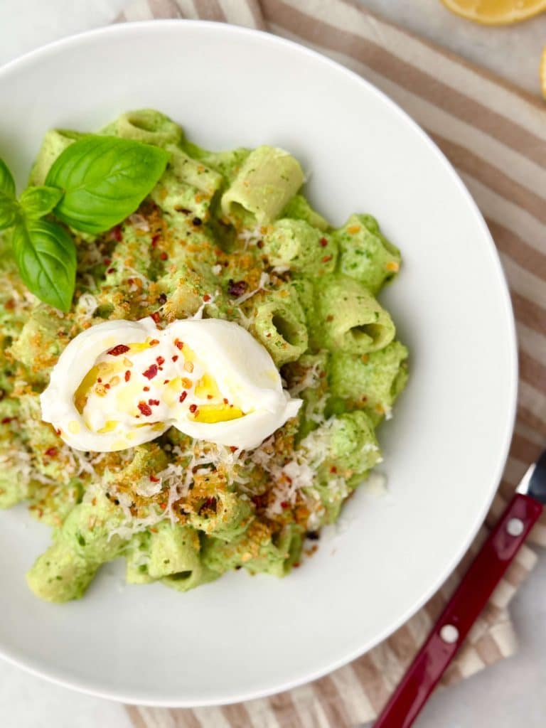 Overhead view of green pea pasta in a white bowl with fresh basil, crispy breadcrumbs, and a ball of burrata.