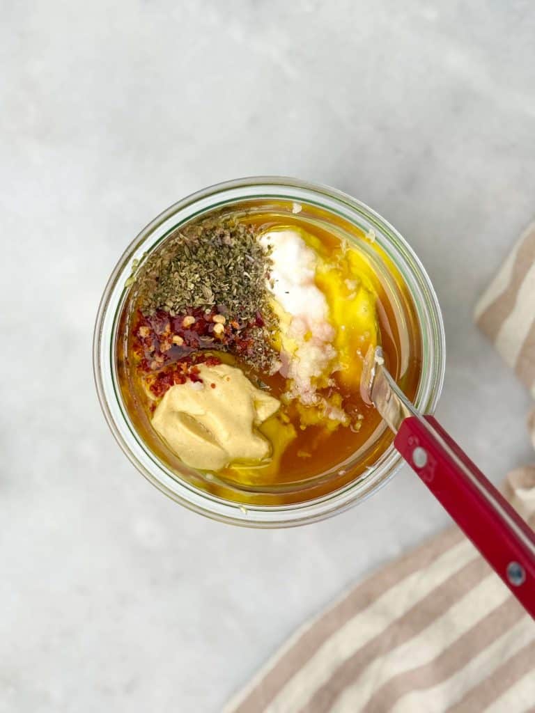 Overhead shot of creamy grinder bean salad dressing ingredients including Greek yogurt, Dijon mustard, olive oil, oregano, and red pepper flakes in a glass jar.
