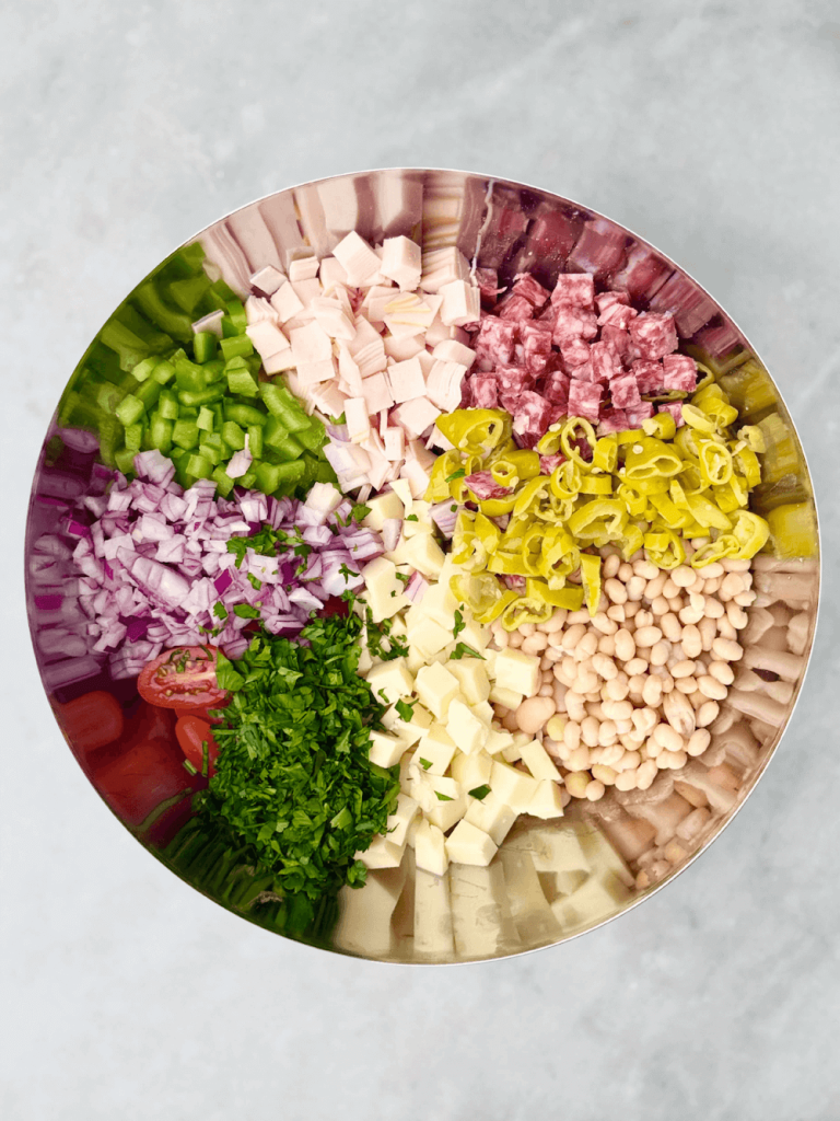 Large mixing bowl filled with colorful ingredients for Italian grinder bean salad before tossing, including beans, salami, turkey, cheese, and vegetables.