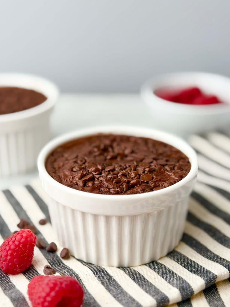 Chocolate high protein baked oats with Greek yogurt in a white ramekin topped with mini chocolate chips, served on a striped cloth with fresh raspberries.