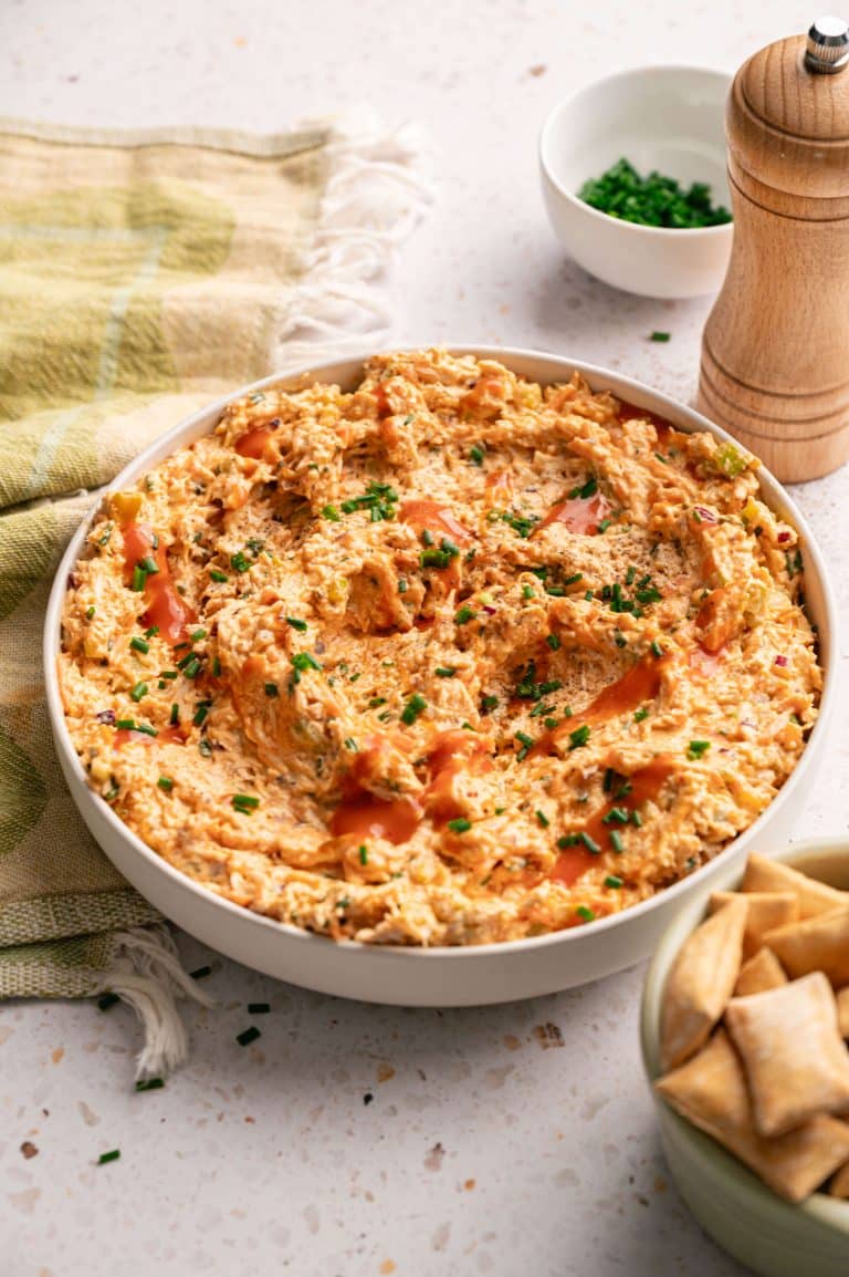 Bowl of buffalo chicken salad drizzled with hot sauce and topped with fresh chives, served with pita chips on the side.