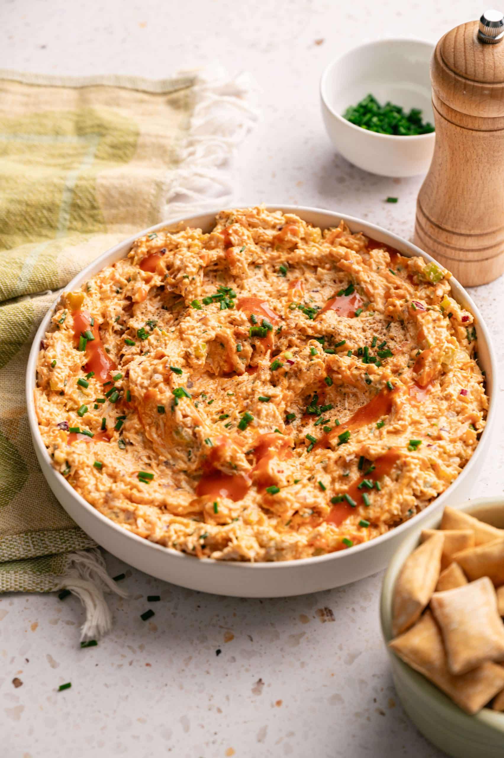 Bowl of buffalo chicken salad drizzled with hot sauce and topped with fresh chives, served with pita chips on the side.