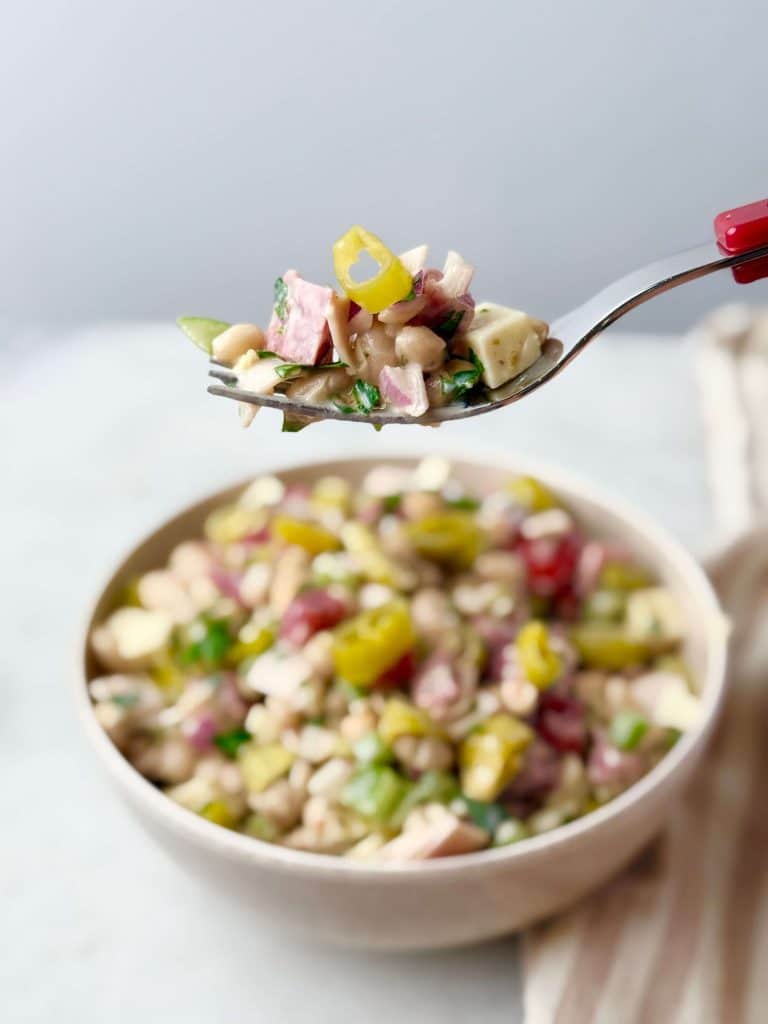 Fork lifting a bite of Italian grinder bean salad with beans, diced deli meats, cheese, and vegetables over a bowl of salad on a light background.