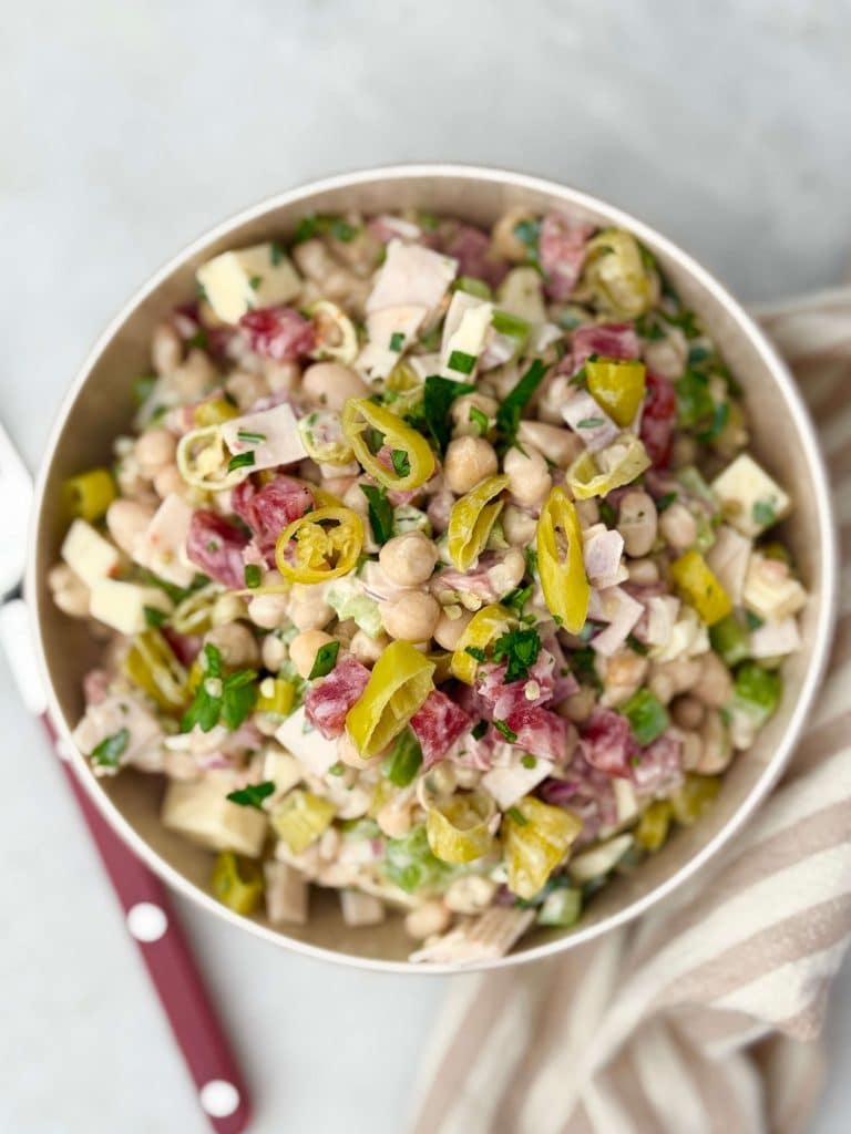 Italian grinder bean salad in a white bowl with diced turkey, salami, mozzarella, chickpeas, white beans, pepperoncini, and fresh parsley on a light background.