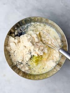 Mixing bowl filled with oats, Greek yogurt, milk, lemon zest, and protein powder for lemon poppy seed overnight oats, with a metal whisk resting on the side