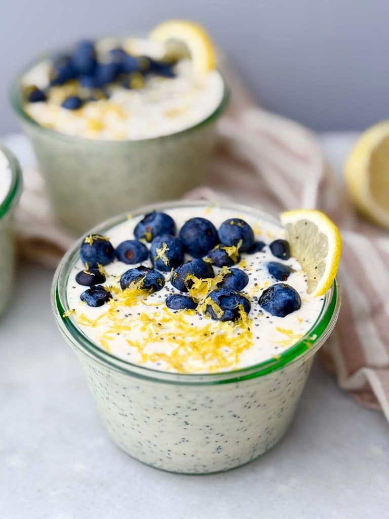 Side view of glass jars filled with lemon poppy seed overnight oats, topped with blueberries and lemon zest, with a striped napkin and lemon halves in the background
