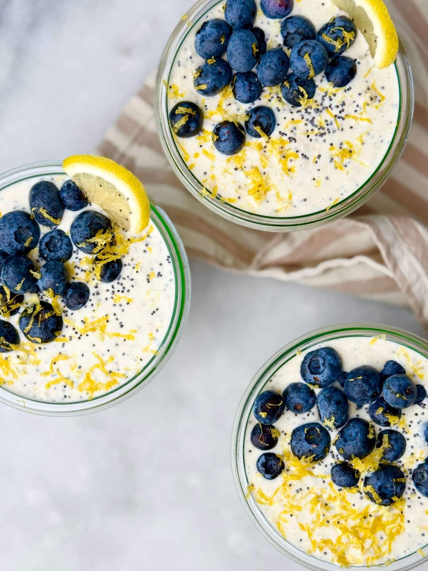 Top-down view of three glass jars filled with lemon poppy seed overnight oats, topped with fresh blueberries, lemon zest, and a lemon wedge on a white background