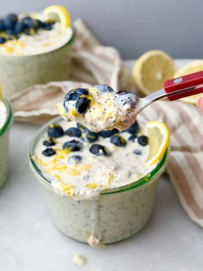 Spoonful of creamy lemon poppy seed overnight oats lifted over a glass jar, showing blueberries, poppy seeds, and lemon zest, with more jars and lemons in the background