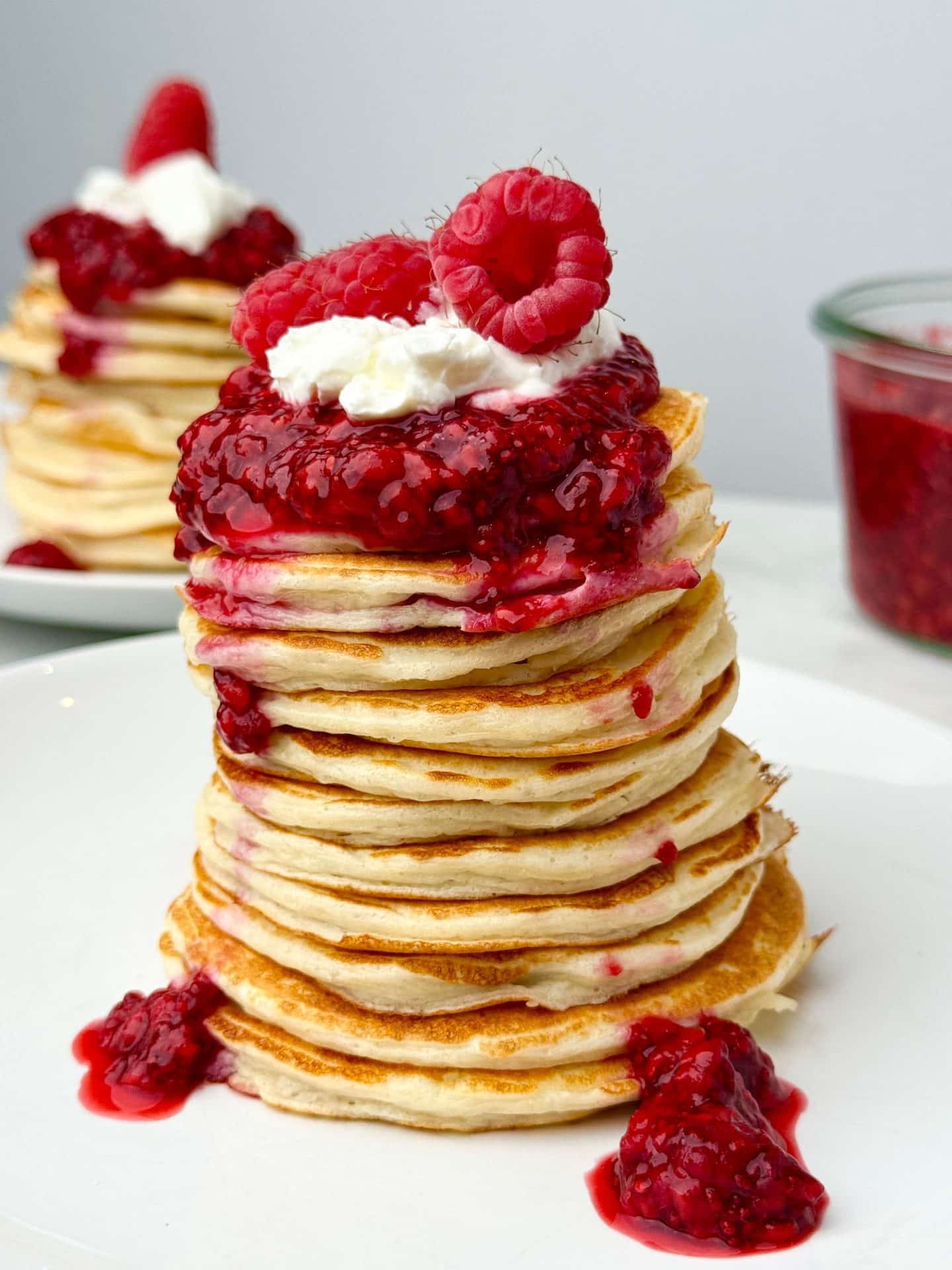 Tall stack of low calorie protein pancakes topped with raspberry chia jam and fresh raspberries on a white plate, photographed in bright natural light.