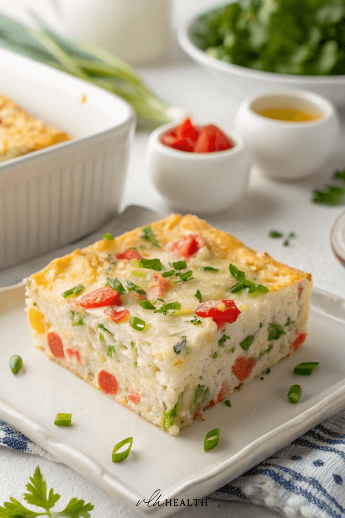 Square slice of egg bake with shredded potatoes, bell peppers, and herbs, served on a white plate with fresh chopped green onions.