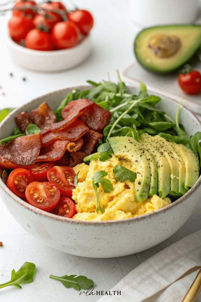 Bowl filled with scrambled eggs, sliced avocado, crispy bacon, cherry tomatoes, and fresh greens on a white background.