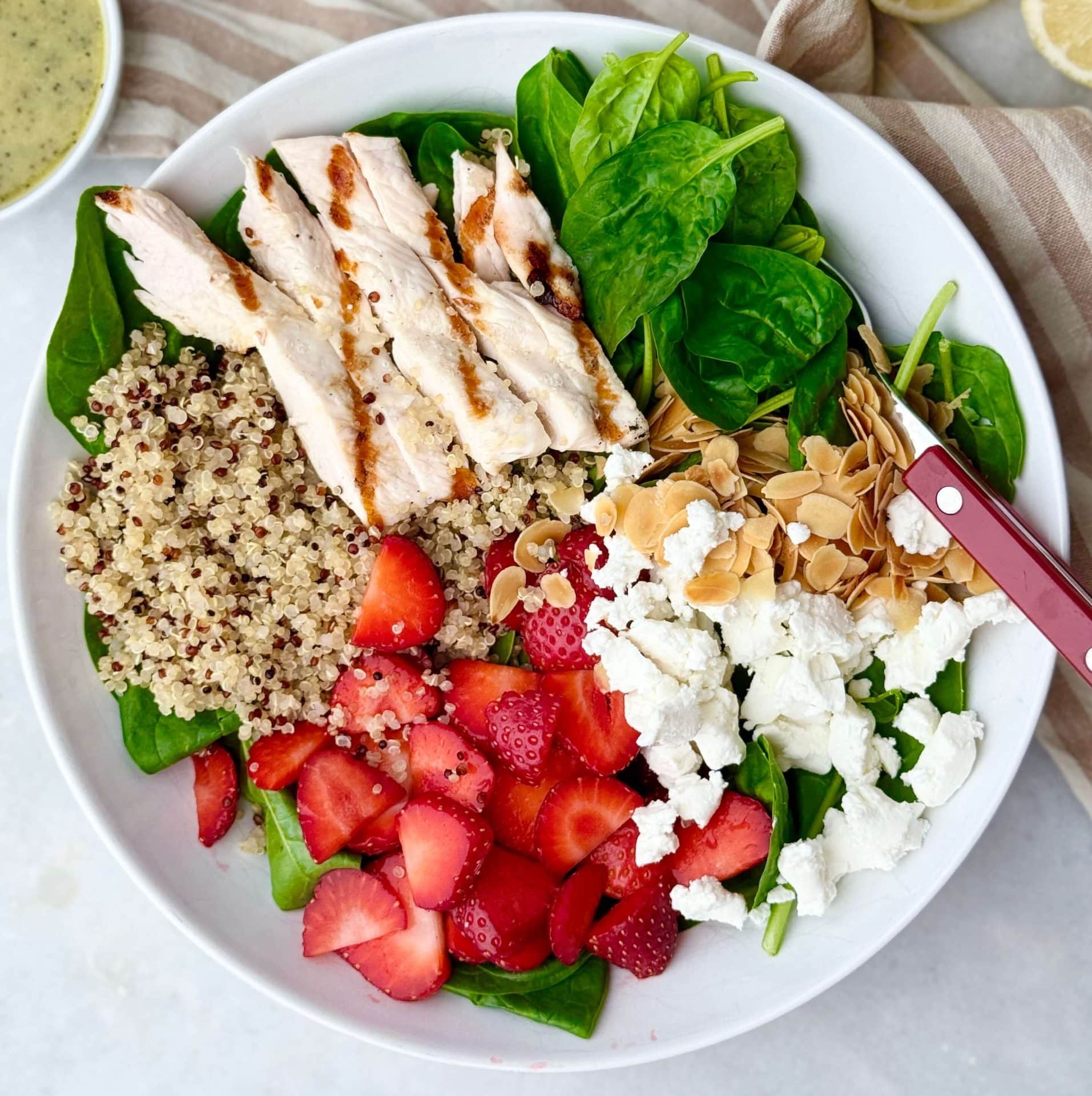 Overhead view of spinach strawberry salad with quinoa, grilled chicken, fresh strawberries, and goat cheese in a white bowl.