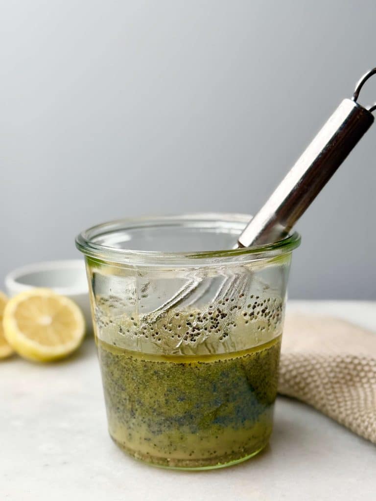Homemade poppyseed dressing in a glass jar with a whisk, surrounded by fresh lemon halves.