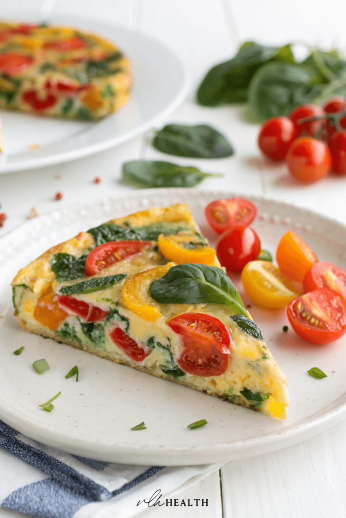 Wedge of colorful vegetable frittata with spinach, cherry tomatoes, and bell peppers on a white plate.