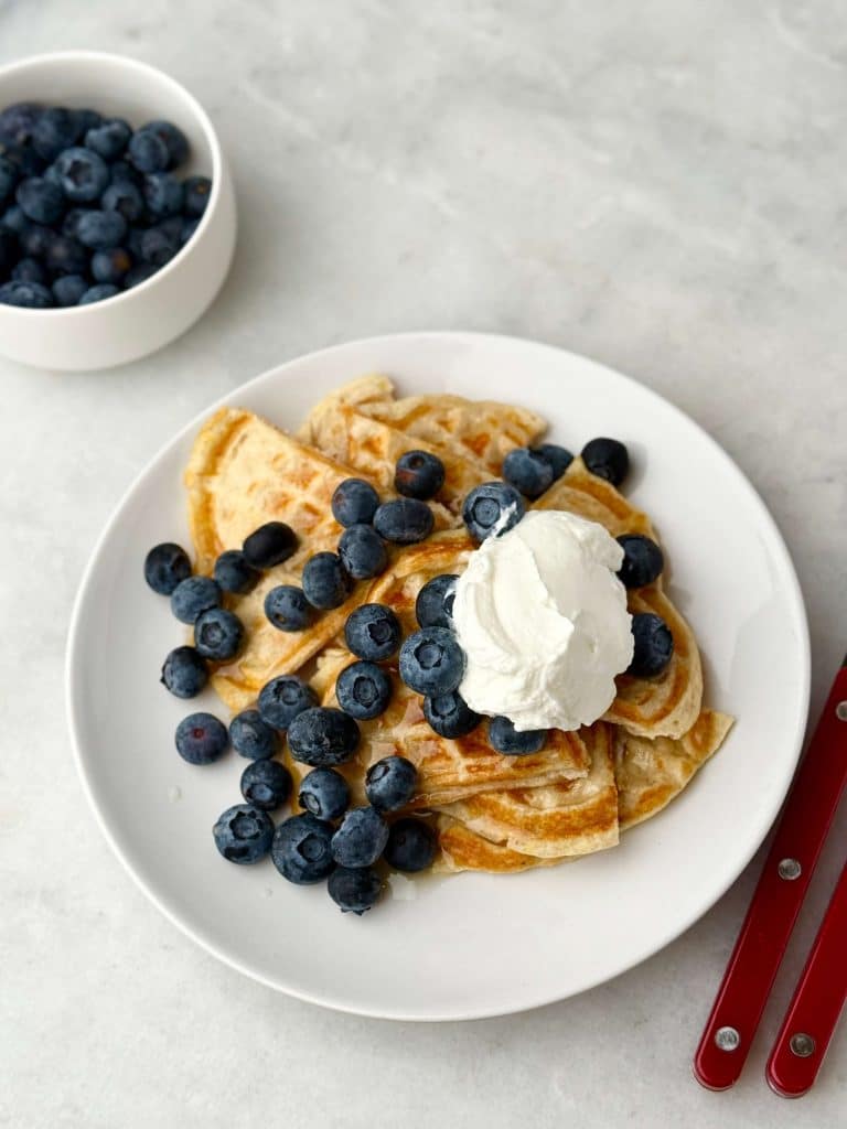 Close-up of fluffy 3 ingredient protein waffles topped with fresh blueberries, a dollop of Greek yogurt, and a drizzle of syrup on a white plate, with extra blueberries in a small bowl in the background.