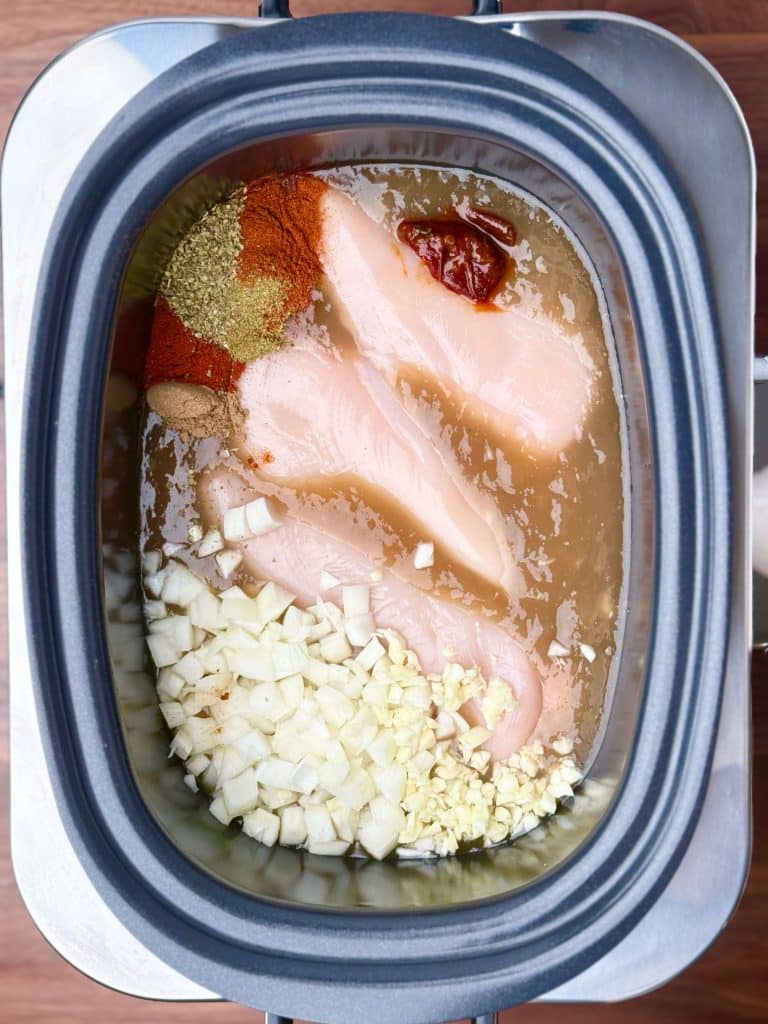 Raw chicken breasts, onions, garlic, chipotle, and spices in a slow cooker with bone broth before cooking.