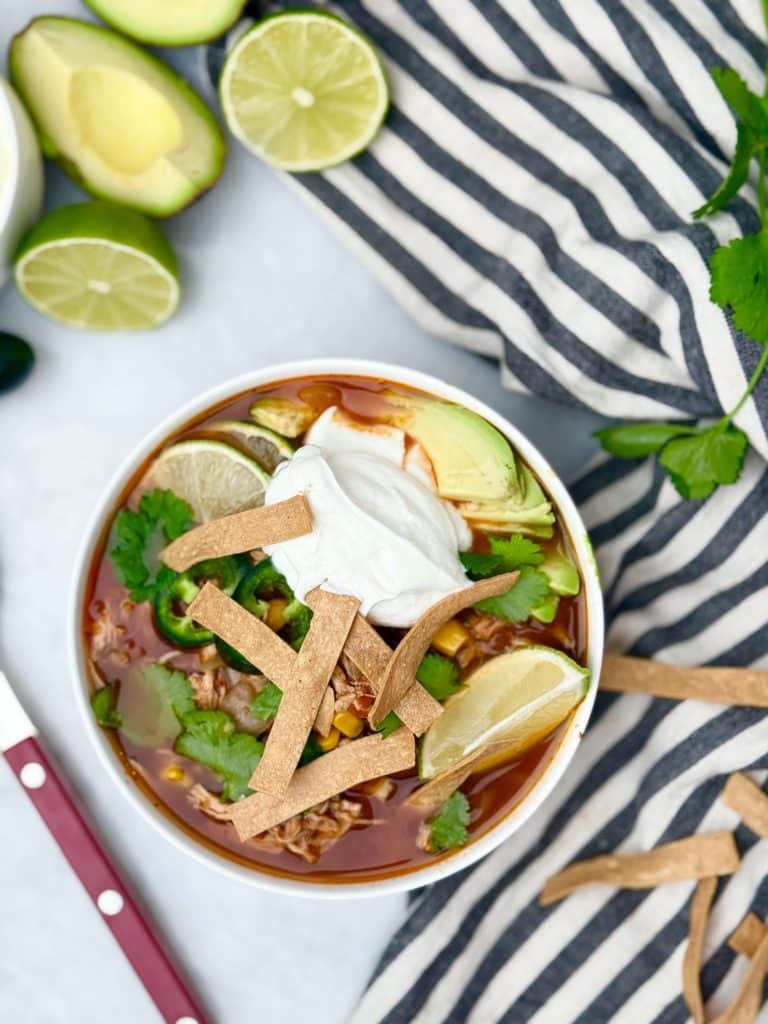 Crockpot chicken tortilla soup served with creamy yogurt, fresh cilantro, avocado, lime wedges, and crunchy tortilla strips.