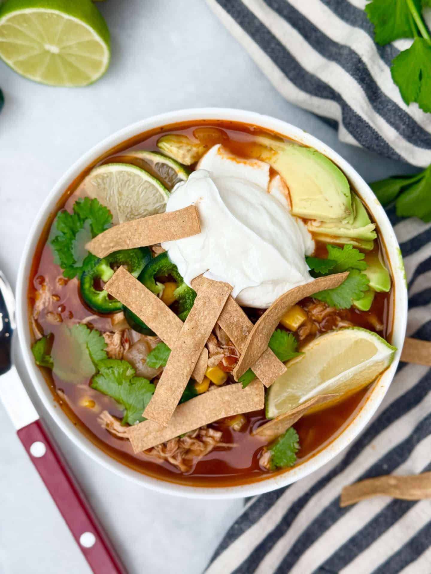 Overhead view of a bowl of crockpot chicken tortilla soup topped with Greek yogurt, avocado slices, cilantro, jalapeño, lime wedge, and baked tortilla strips.