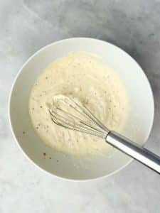Creamy Greek yogurt and mayo dressing with mustard and pickle juice whisked in a white bowl on marble countertop.