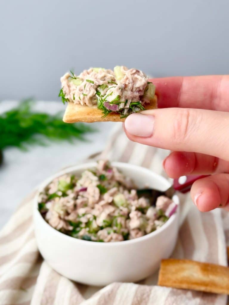 Close‑up of a cracker topped with healthy dill pickle tuna salad, held over a bowl of tuna salad on striped linen.