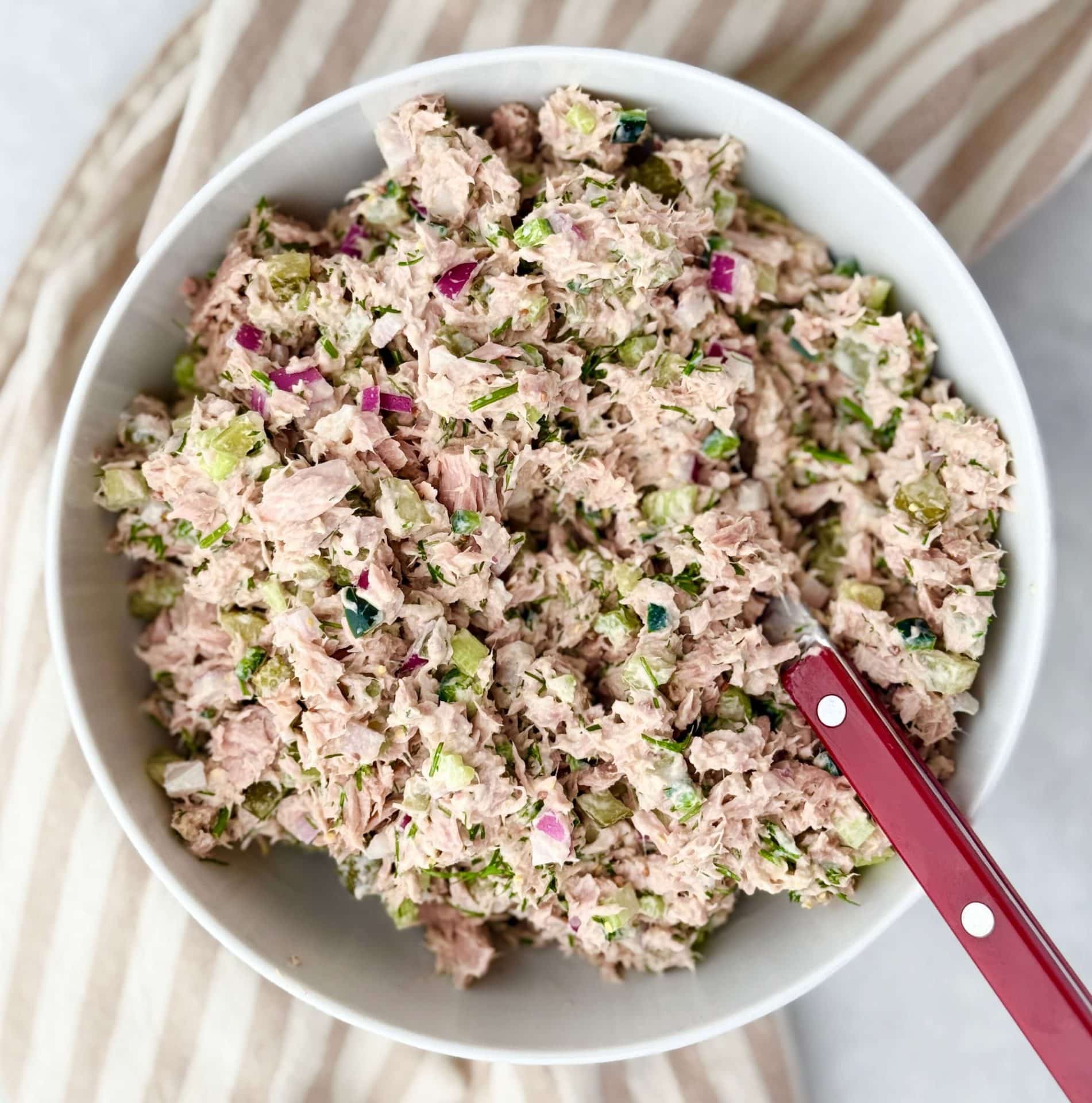 Healthy dill pickle tuna salad in white bowl with red spoon, surrounded by striped fabric for a casual meal prep look.
