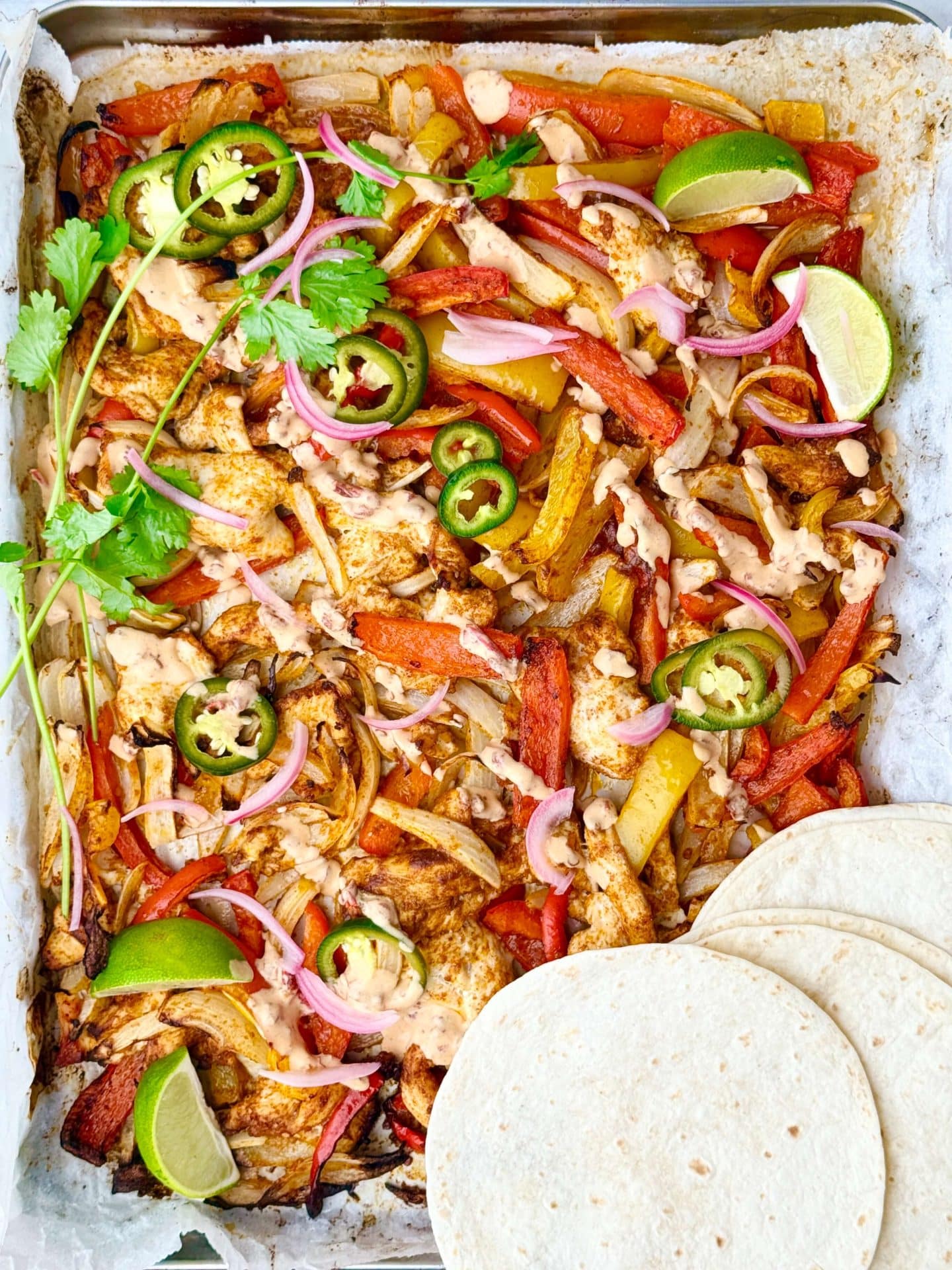 Colorful sheet pan chicken fajitas with roasted bell peppers and onions, drizzled with chipotle crema and topped with pickled jalapeños, pickled red onions, cilantro, and lime wedges, served with warm tortillas.