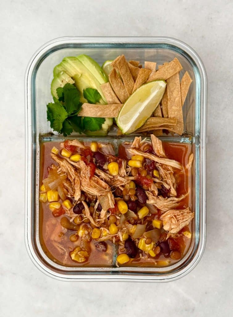 Glass meal prep container with a portion of chicken tortilla soup, avocado slices, cilantro, lime, and crispy tortilla strips for an easy high-protein lunch.