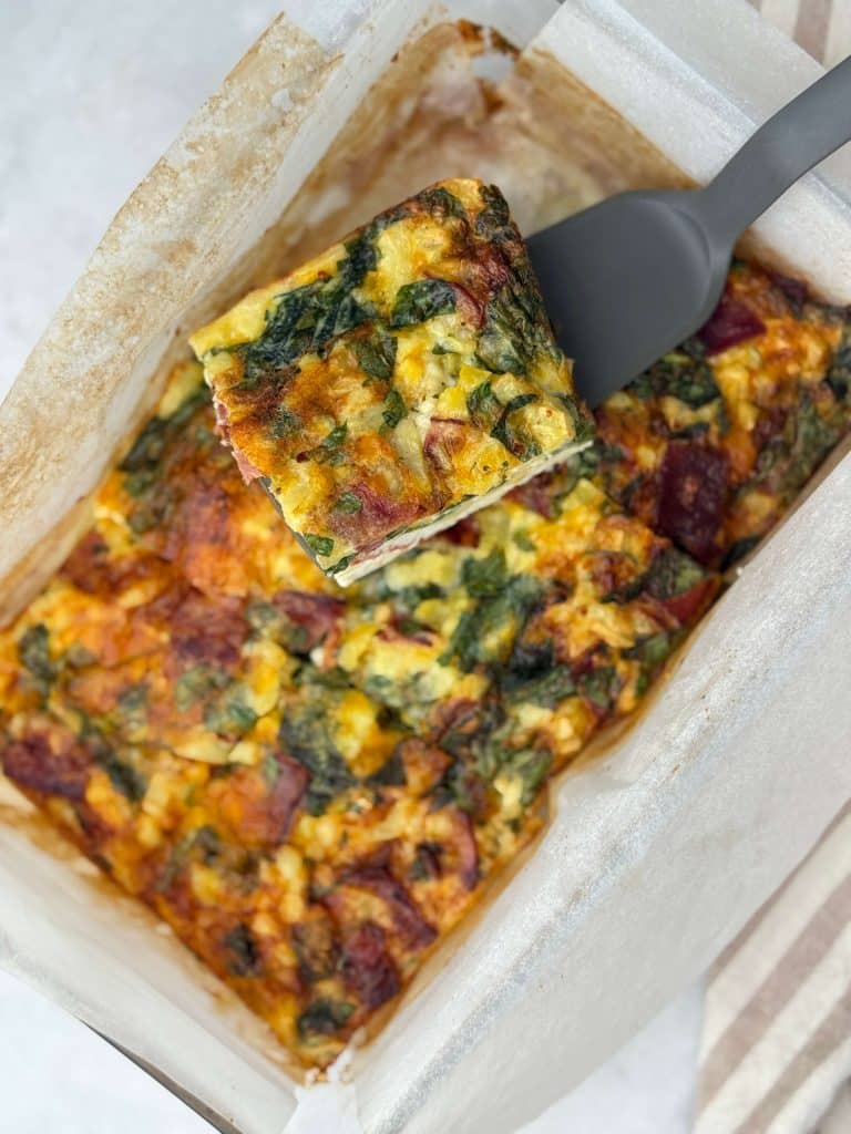 Overhead view of baked high protein egg casserole with hash browns, spinach, cheddar cheese, and turkey bacon in a glass dish lined with parchment.