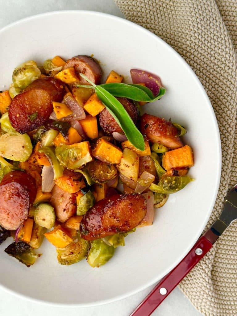 Plated fall sausage and veggie sheet pan dinner with maple mustard butter and fresh sage garnish.