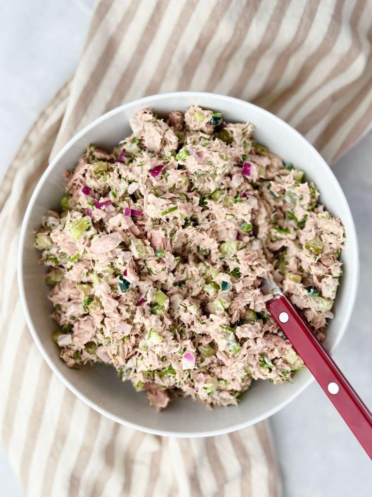 Overhead shot of healthy dill pickle tuna salad in a white bowl on a striped napkin, mixed with fresh herbs and crunchy vegetables.