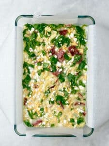 Unbaked high protein egg bake mixture with cottage cheese, spinach, hash browns, cheddar cheese, and turkey bacon in a parchment-lined baking dish.