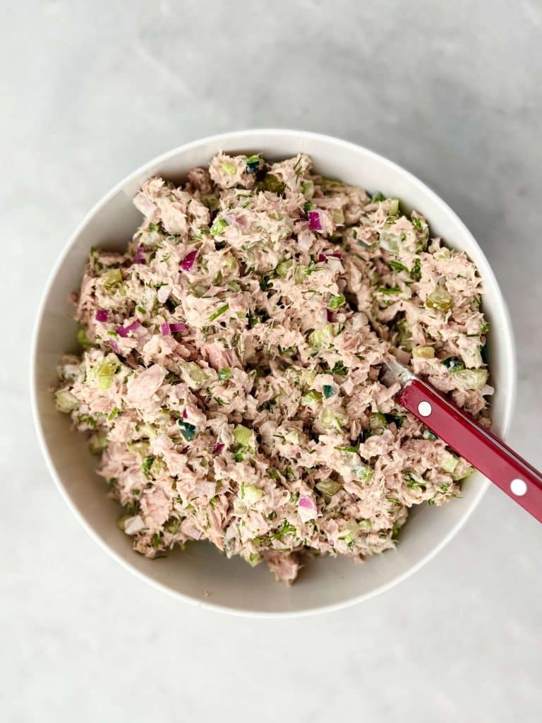 Close‑up of healthy dill pickle tuna salad mixed in a white bowl with red spoon, showing celery, pickles, and fresh dill.