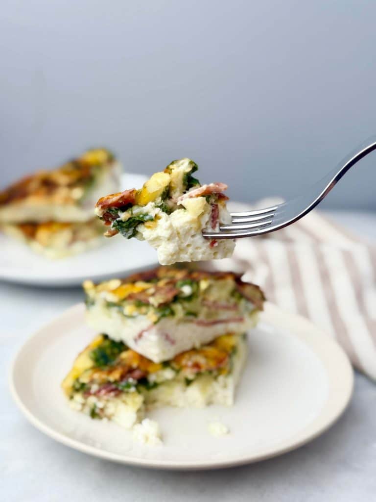 Fork holding a bite of high protein cottage cheese egg bake with spinach and turkey bacon, served on a plate stacked with casserole bars for breakfast.