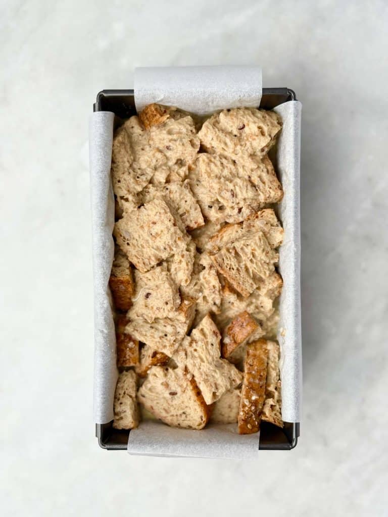 Cubes of high protein bread soaked in custard mixture arranged in a parchment-lined loaf pan before baking.