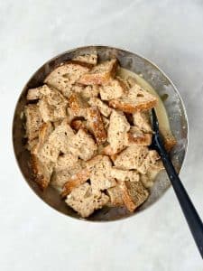 Cubes of high protein bread soaking in the custard mixture in a stainless steel bowl before baking.