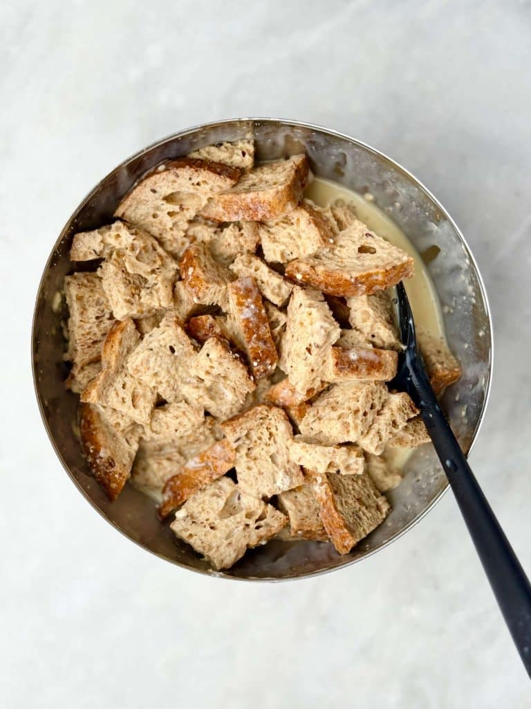 Cubes of high protein bread soaking in the custard mixture in a stainless steel bowl before baking.