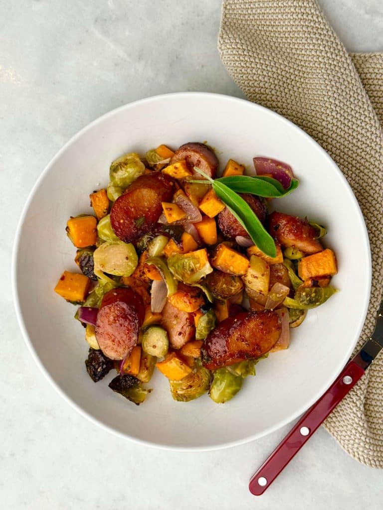 Fall sausage and veggie sheet pan dinner served in a white bowl with sage garnish.